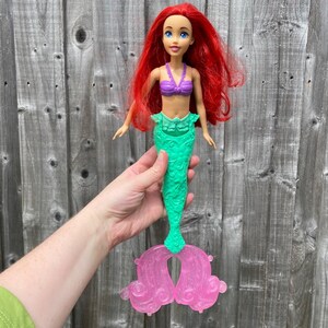 May include: A doll of a mermaid with red hair, blue eyes, and a purple top. The mermaid has a green tail and pink fins. The doll is held in a person's hand against a wooden background.
