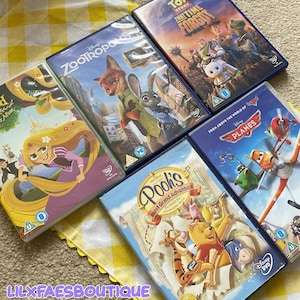 Puede incluir: Una colección de DVDs de Disney, incluyendo "Enredados," "Zootrópolis," "Toy Story That Time Forgot," "Aviones" y "Las Grandes Aventuras de Winnie the Pooh." Los DVDs están en sus estuches originales, sobre una superficie a cuadros amarillos y blancos. El texto "LILXFAESBOUTIQUE" es visible.