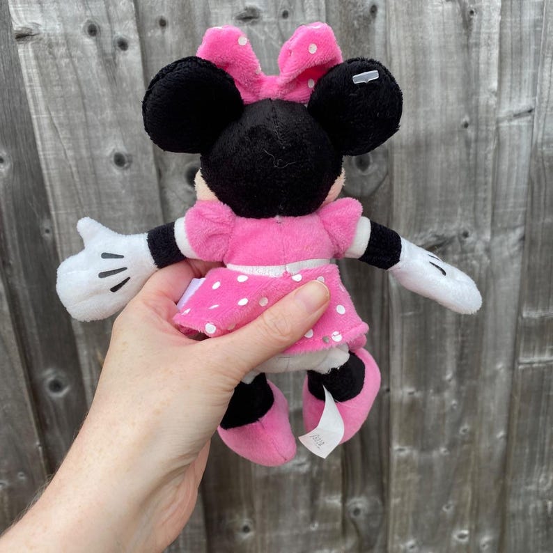 May include: A Minnie Mouse plush toy. The plush features black ears, a pink bow with white polka dots, and a pink dress with white polka dots. The toy has white gloves and black shoes. The toy is held in a hand.
