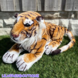Op de afbeelding: Een pluche tijgerknuffel met oranje en zwarte strepen, witte vacht op de snuit en borst, donkerbruine oren en zwarte plastic ogen. Het knuffeldier ligt in een liggende houding met de voorpoten naar voren uitgestrekt en een lange gestreepte staart.