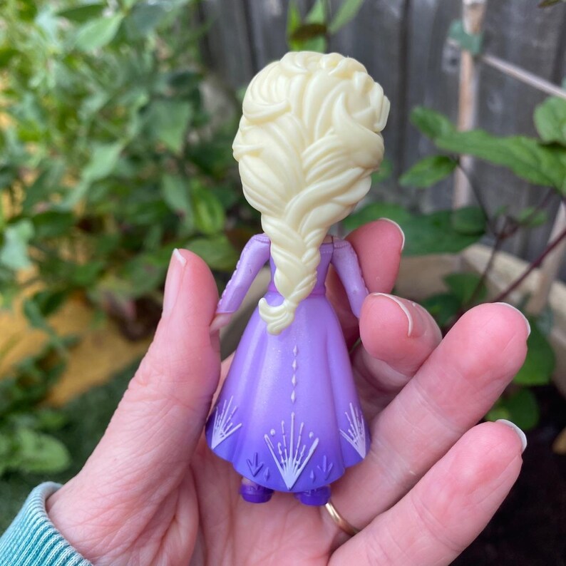 Puede incluir: Una peque&ntilde;a figura de pl&aacute;stico que representa a un personaje con cabello largo de color rubio p&aacute;lido peinado en una gruesa trenza. La figura viste un vestido morado que se oscurece hacia el dobladillo, decorado con patrones de copos de nieve blancos.
