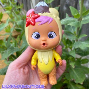 May include: A doll with a yellow ice cream cone-shaped body and a matching hood. The doll has a surprised expression, large eyes, and pink and purple hair. A red flower and a chocolate and vanilla ice cream cone top the hood. The text "LILXFAESBOUTIQUE" is at the bottom.