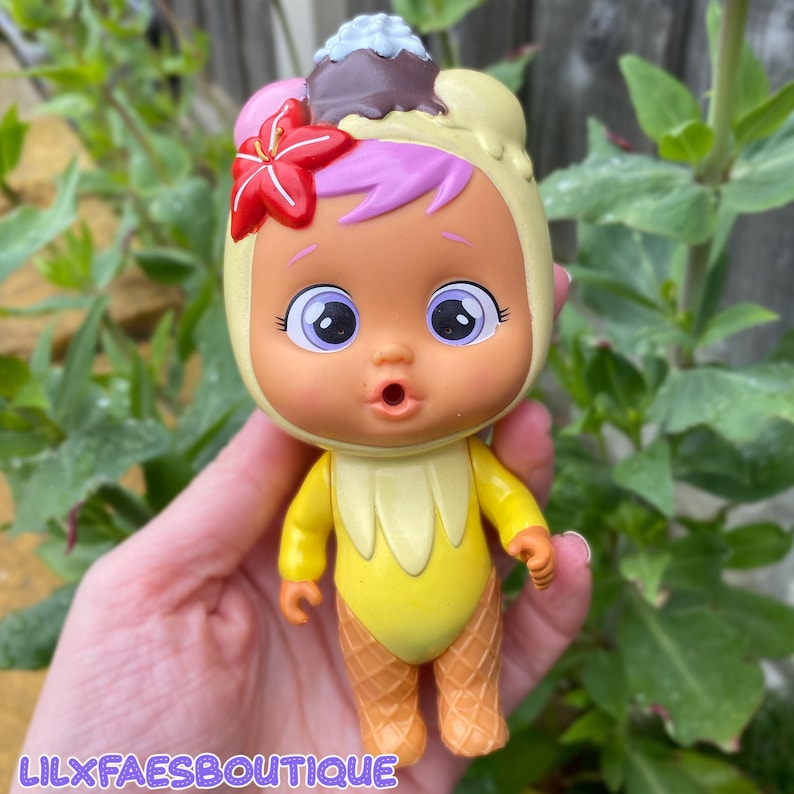 May include: A doll with a yellow ice cream cone-shaped body and a matching hood. The doll has a surprised expression, large eyes, and pink and purple hair. A red flower and a chocolate and vanilla ice cream cone top the hood. The text "LILXFAESBOUTIQUE" is at the bottom.