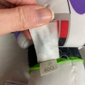 K&ouml;nnte beinhalten: Nahaufnahme eines wei&szlig;en Etiketts mit der Aufschrift "69" und "40G". Das Etikett ist an einem wei&szlig;en und schwarzen Stofftier mit lila Akzenten befestigt. Das Etikett hat auch Text, der teilweise sichtbar ist.
