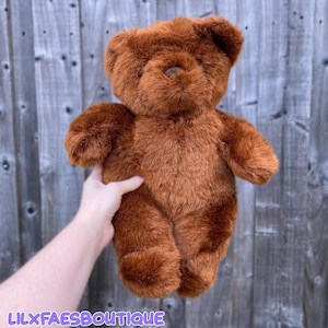 Puede incluir: Un oso de peluche marrón con una textura suave y afelpada. El oso tiene una cabeza redonda, orejas pequeñas y una nariz de botón. El oso está siendo sostenido por una mano. El texto "LILXFAESBOUTIQUE" está en la parte inferior de la imagen.
