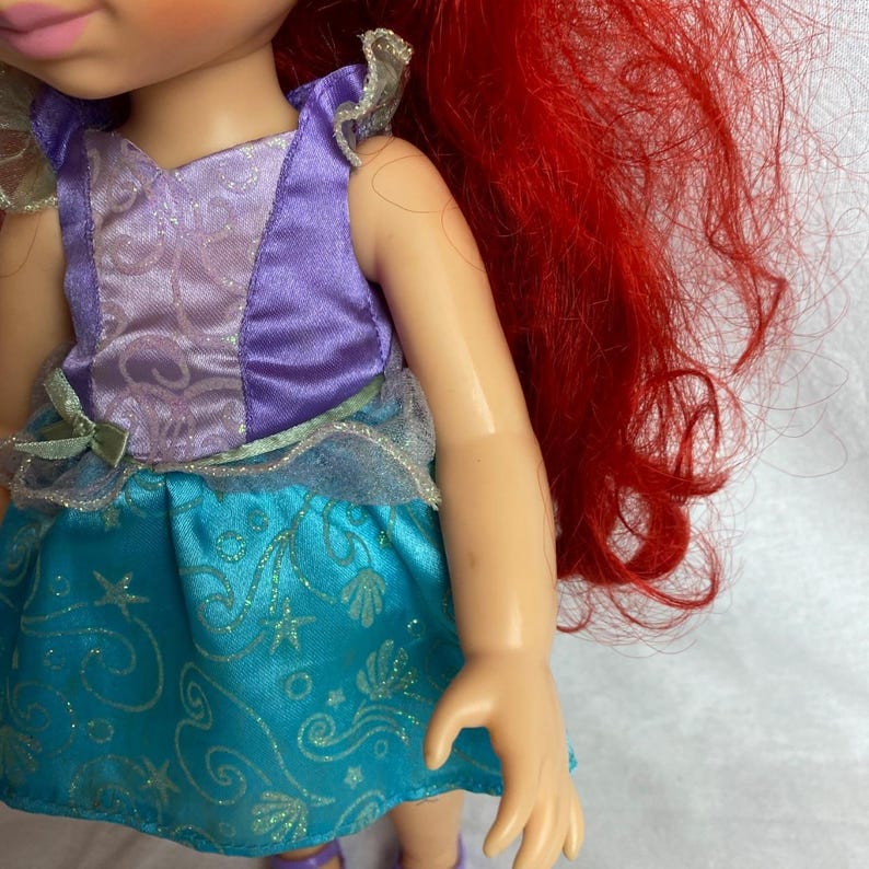 Puede incluir: Una mu&ntilde;eca con cabello rojo y un vestido morado y turquesa. El vestido de la mu&ntilde;eca tiene una falda turquesa con un estampado de conchas marinas y estrellas. La parte superior del vestido es morada con detalles plateados. El cabello de la mu&ntilde;eca es largo y ondulado.