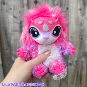 Peut inclure: Une peluche créature avec une fourrure rose vif, de grands yeux expressifs et un nez violet clair. Le jouet présente un ventre en fourrure blanche, des pattes en plastique rose avec des coussinets moulés et des petites cornes sur le front.