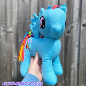 Könnte beinhalten: Ein hellblaues Plüsch-Pony mit einer regenbogenfarbenen Mähne und einem Schweif, großen gestickten Augen und einem Wolkensymbol mit einem Regenbogen-Blitz auf der Flanke. Das Spielzeug hat kleine Flügel am Rücken und eine rote Haartolle am Kopf.