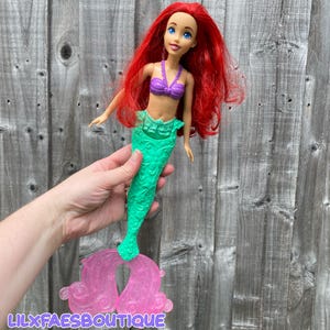 May include: A doll of a mermaid with long red hair, blue eyes, and a purple top. The mermaid has a green tail and a pink stand. The doll is held in front of a wooden background. The text "LILXFAESBOUTIQUE" is visible at the bottom.