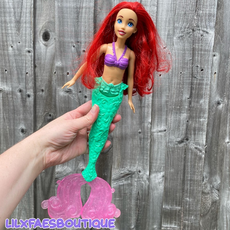 May include: A doll of a mermaid with long red hair, blue eyes, and a purple top. The mermaid has a green tail and a pink stand. The doll is held in front of a wooden background. The text "LILXFAESBOUTIQUE" is visible at the bottom.
