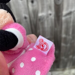May include: A pink plush toy with white polka dots and a black face. The toy has a small tag with a red logo. The toy is held in a person's hand.
