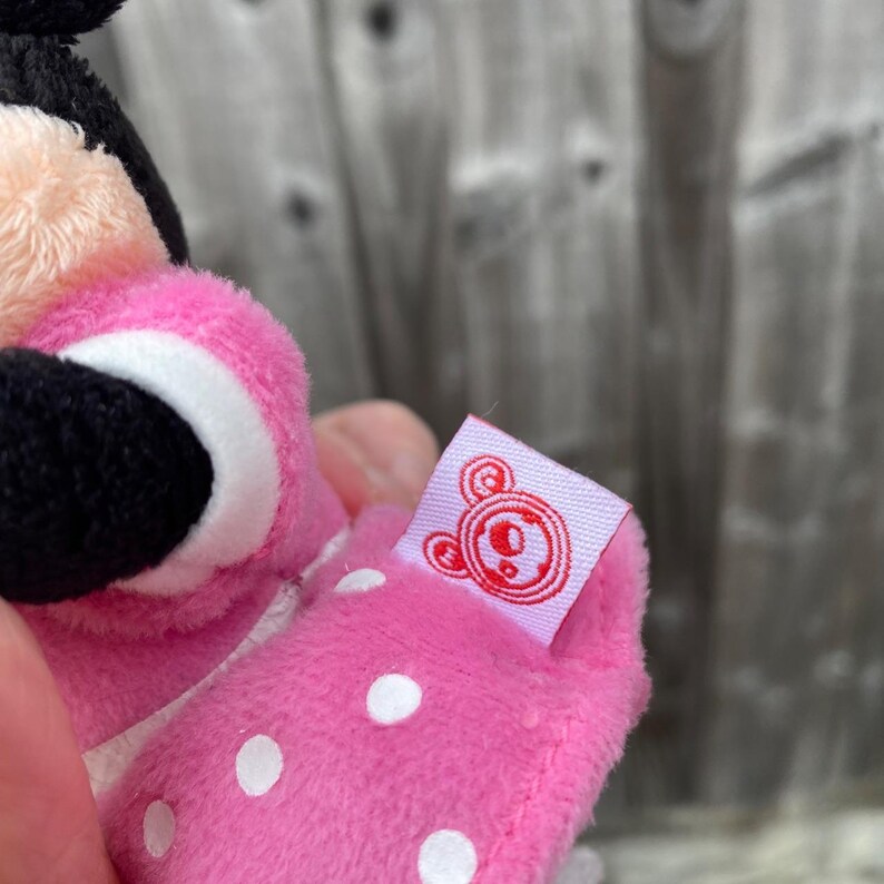 May include: A pink plush toy with white polka dots and a black face. The toy has a small tag with a red logo. The toy is held in a person's hand.