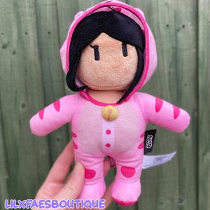 May include: A plush toy character wearing a pink hooded onesie with dark pink tiger-like stripes. The character has black hair, a tan face with simple embroidered features, and a small yellow bell detail on the chest. A black tag reads Stumble Guys.