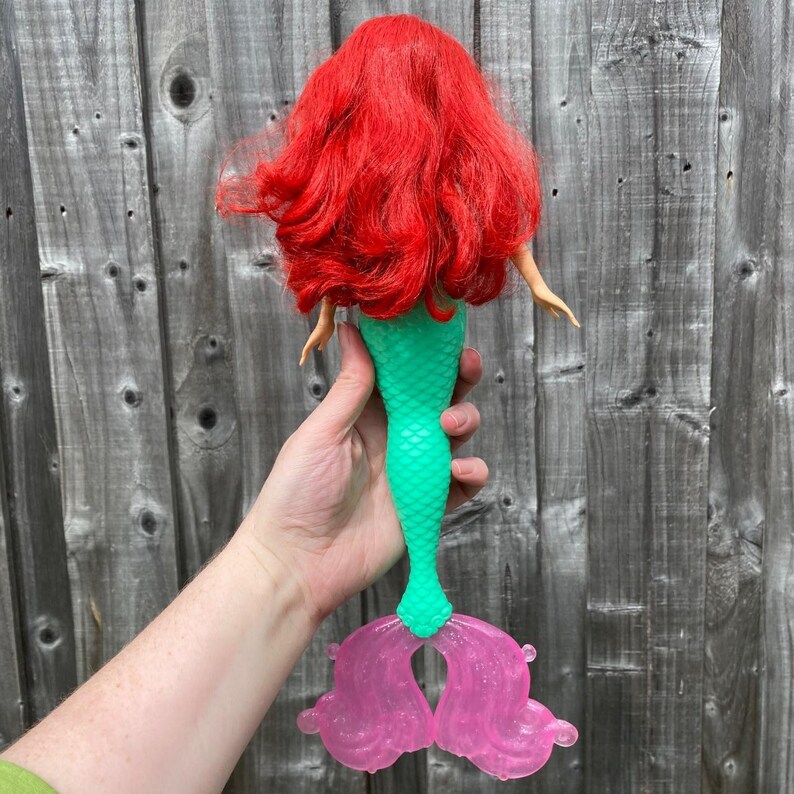 May include: A doll with bright red hair and a green mermaid tail. The tail has a pink, translucent fin. The doll is held up against a weathered wooden fence.