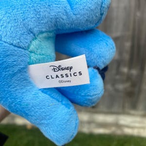 May include: A blue plush toy with a white label displaying "Disney CLASSICS" and the Disney logo. The toy is soft and fluffy, with visible stitching and a textured surface.