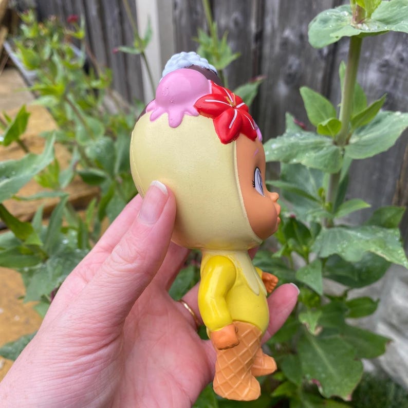 May include: A yellow figurine of a cartoon character wearing a yellow outfit with waffle cone feet. The character has a light yellow head covering with a pink, red, and brown ice cream cone on top. The figurine is held in a hand.