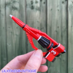 Peut inclure: Un stylo à bille en plastique rouge en forme de fusée Thunderbird 3. Le stylo présente des détails de moteur noirs, un chiffre 3 blanc sur le côté et le texte THUNDER BIRD imprimé en blanc le long de la section conique du nez de la fusée.