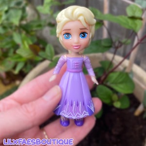 Puede incluir: Una peque&ntilde;a mu&ntilde;eca de pl&aacute;stico con cabello rubio recogido, ojos azules grandes y tez clara. La mu&ntilde;eca viste un vestido morado de manga larga con una falda de color morado m&aacute;s oscuro que presenta patrones geom&eacute;tricos blancos. El texto LILXFAESBOUTIQUE es visible.