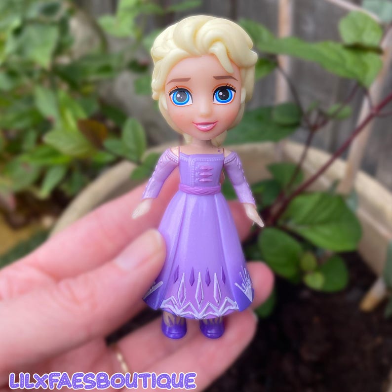 Puede incluir: Una peque&ntilde;a mu&ntilde;eca de pl&aacute;stico con cabello rubio recogido, ojos azules grandes y tez clara. La mu&ntilde;eca viste un vestido morado de manga larga con una falda de color morado m&aacute;s oscuro que presenta patrones geom&eacute;tricos blancos. El texto LILXFAESBOUTIQUE es visible.