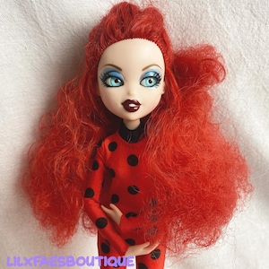 Könnte beinhalten: Eine Puppe mit leuchtend rotem, lockigem Haar und blauem Lidschatten. Die Puppe trägt einen roten Overall mit schwarzen Punkten. Die Puppe hat dunkelroten Lippenstift und der Text "LILXFAESBOUTIQUE" ist unten sichtbar.