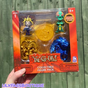 Peut inclure: Un pack de figurines de collection Yu-Gi-Oh! contenant quatre petites figurines : un personnage aux cheveux en pointe, une créature verte, une figure dorée rappelant un pharaon et un dragon bleu. La boîte inclut un emblème doré et le logo Yu-Gi-Oh!.