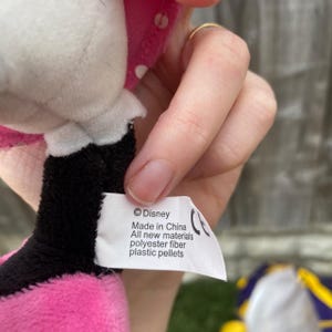May include: A close-up of a Minnie Mouse plush toy. The toy is pink, black, and white. A white tag with the Disney logo and text is visible. The tag reads "Made in China, All new materials, polyester fiber, plastic pellets."