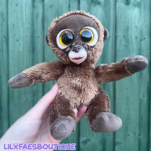 Puede incluir: Un peluche de mono con pelaje marrón suave, grandes ojos dorados brillantes y una cara de color marrón claro. El animal de peluche presenta una nariz pequeña y oscura y una boca cosida. El texto LILXFAESBOUTIQUE está impreso en morado abajo.