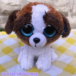 May include: A plush toy dog with brown, white, and tan fur, and large blue glittery eyes. The toy dog is sitting on a yellow and white checkered surface. The text "LILXFAESBOUTIQUE" is at the bottom of the image.