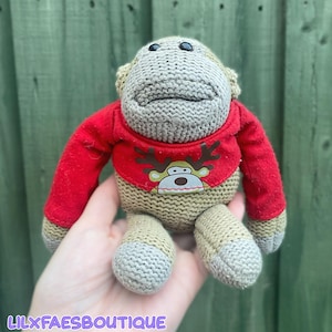 May include: A knitted monkey plush toy featuring a light brown body and a red long-sleeved shirt. The shirt displays a graphic of a reindeer face with brown antlers. The monkey has black bead eyes and a textured, hand-knit appearance throughout.