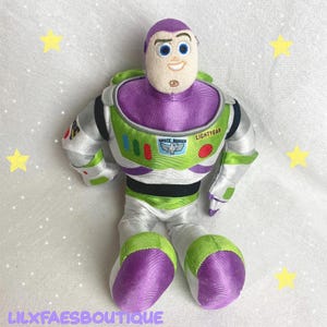 May include: A plush Buzz Lightyear doll, featuring a purple head and accents, a green and white spacesuit, and the words "Space Ranger" and "Lightyear" on the chest. The doll is sitting with its legs extended. The background includes yellow star decorations.