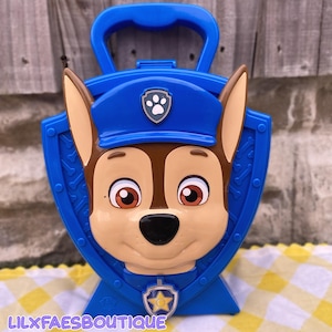 May include: Blue plastic Chase the police dog carrying case with a handle. The case features Chase's face, a blue police hat, and a badge. The text "LILXFAESBOUTIQUE" is at the bottom.