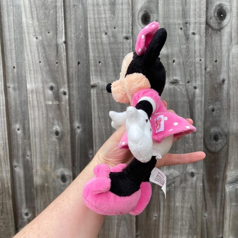 May include: A Minnie Mouse plush toy in a side profile. The plush has a pink and white polka dot dress and bow, black ears, and white gloves. The toy is held up against a wooden background.