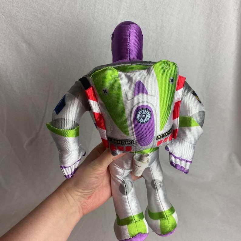 May include: A plush Buzz Lightyear doll, viewed from the back, featuring a purple helmet, white and silver spacesuit, and green and white accents. The doll has a backpack with the text "JET EX" and "DANGER!"