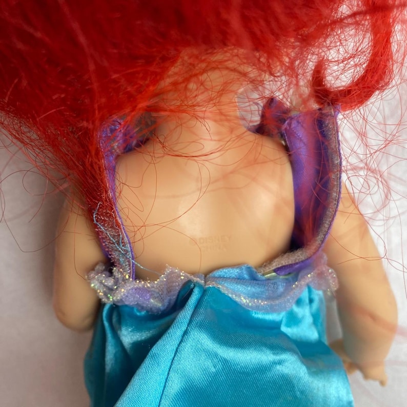 Puede incluir: Mu&ntilde;eca con pelo rojo y vestido turquesa. El vestido de la mu&ntilde;eca tiene un ribete morado con purpurina. La espalda de la mu&ntilde;eca es visible, con la palabra "Disney" impresa en la espalda.