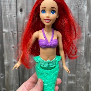 May include: A doll of a mermaid with red hair, blue eyes, and a purple top. The doll has a green mermaid tail. The doll is a toy.