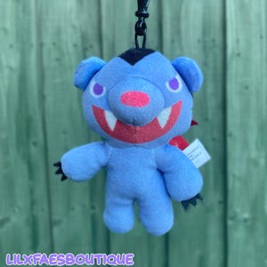 Peut inclure: Une peluche porte-clés bleue représentant une créature stylisée ressemblant à un ours avec une tache noire sur la tête, des yeux violets, un nez rose et une large bouche rouge montrant deux crocs blancs. Elle a des griffes noires et un clip.