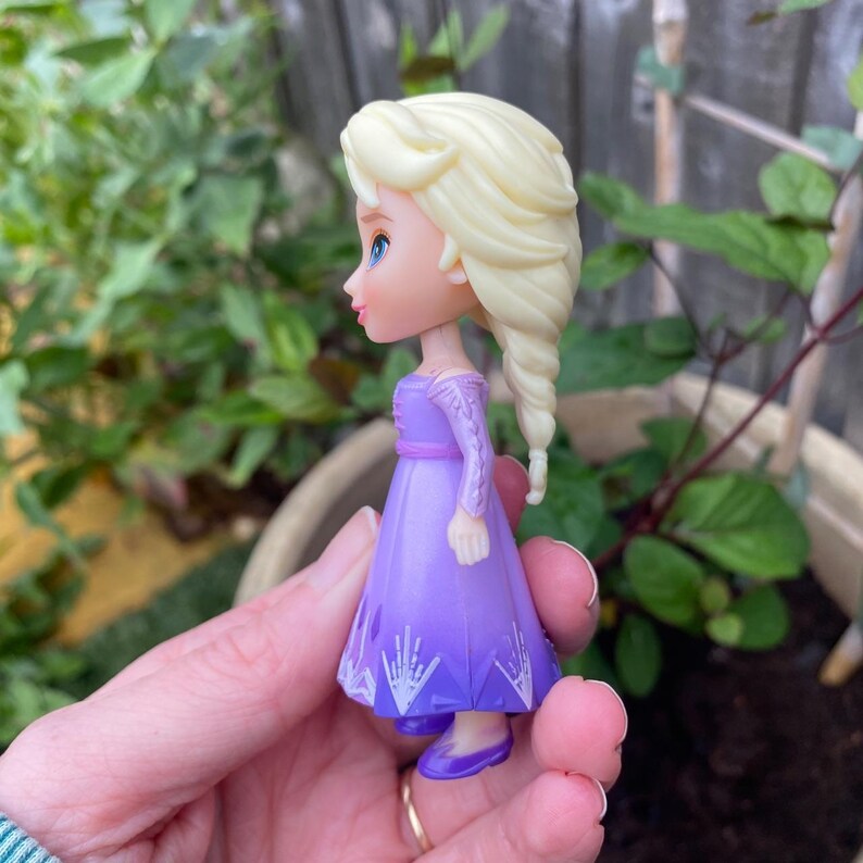Puede incluir: Una peque&ntilde;a figura de pl&aacute;stico de un personaje de piel p&aacute;lida, ojos azules y largo cabello rubio peinado en una trenza lateral. La figura viste un vestido morado de manga larga con un patr&oacute;n de copos de nieve blancos en el dobladillo.