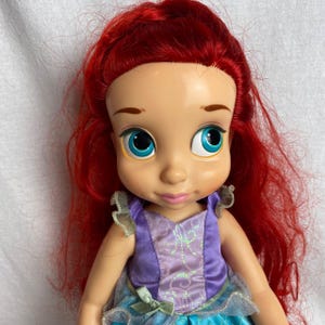 Puede incluir: Mu&ntilde;eca de un personaje con cabello rojo brillante, ojos azules grandes y un vestido morado y azul. La mu&ntilde;eca tiene una tez clara y una sonrisa suave. El cabello de la mu&ntilde;eca est&aacute; peinado en ondas sueltas.