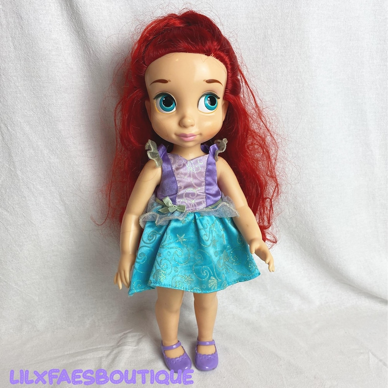 Puede incluir: Mu&ntilde;eca con cabello rojo, ojos azules y tez clara. La mu&ntilde;eca lleva un vestido morado y turquesa y zapatos morados. El texto "LILXFAESBOUTIQUE" est&aacute; en la parte inferior de la imagen.