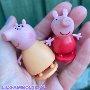May include: Two small Peppa Pig character figurines. One figure is light pink with a peach-coloured dress and a circular base. The second figure is light pink with a red dress and a circular base. Both figures have small arms and ears.