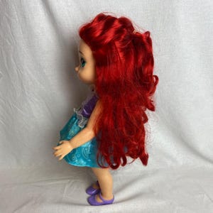 Puede incluir: Mu&ntilde;eca con cabello rojo brillante, vestida con un top morado y un vestido turquesa con detalles blancos. La mu&ntilde;eca tiene ojos azules y zapatos morados. La mu&ntilde;eca est&aacute; de pie y mirando hacia la izquierda.