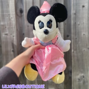 May include: A plush toy of a character with large black ears, a white face, and black nose. The character wears a shiny pink dress with a light blue bodice and a matching pink pointed hat. The text LILXFAESBOUTIQUE is printed in white at the bottom.