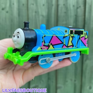 May include: A toy train engine featuring a white face with black eyebrows and eyes, a black smokebox, and a bright blue body decorated with a geometric pattern of pink, yellow, and black shapes. The number 1 is printed in black on the side.