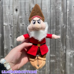 May include: A plush Grumpy dwarf doll from Snow White. The doll has a brown hat with the word "GRUMPY" on it, a white beard, and a red coat with a black belt. The doll is approximately 15 cm tall.