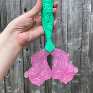 May include: A mermaid tail toy with a green, textured top and translucent pink fins. The fins have a glittery finish and a swirling design. The toy is held against a wooden fence background.