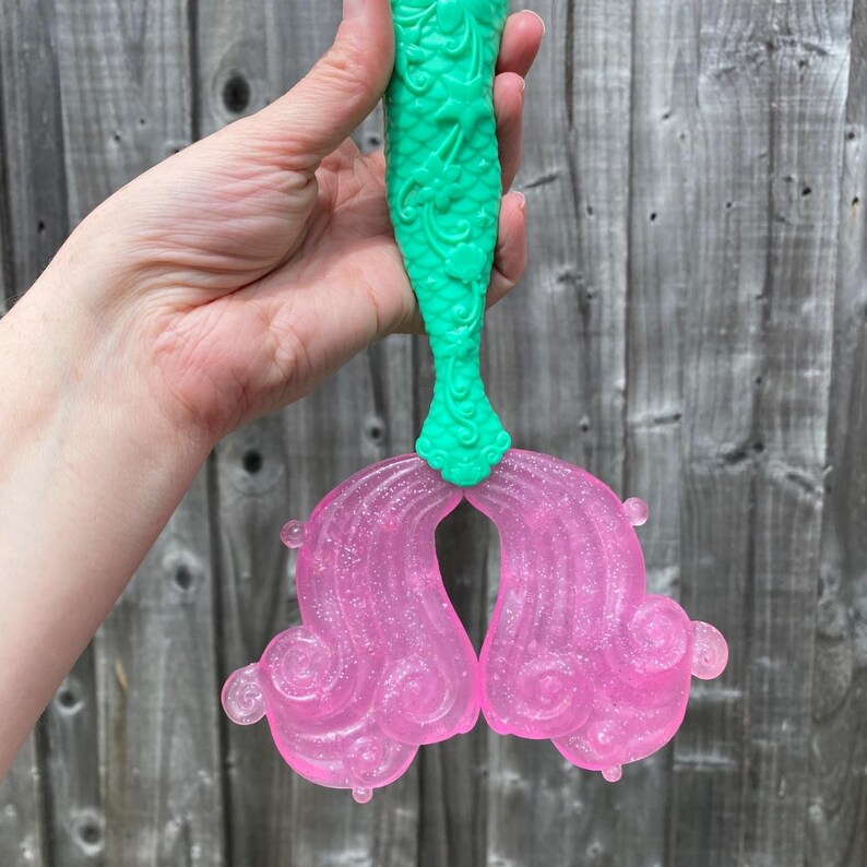 May include: A mermaid tail toy with a green, textured top and translucent pink fins. The fins have a glittery finish and a swirling design. The toy is held against a wooden fence background.