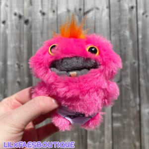 May include: A plush, pink creature with orange hair tufts and large yellow eyes. It has a gray mouth and a single tooth. The creature is wearing a purple diaper. The text "LILXFAESBOUTIQUE" is at the bottom.