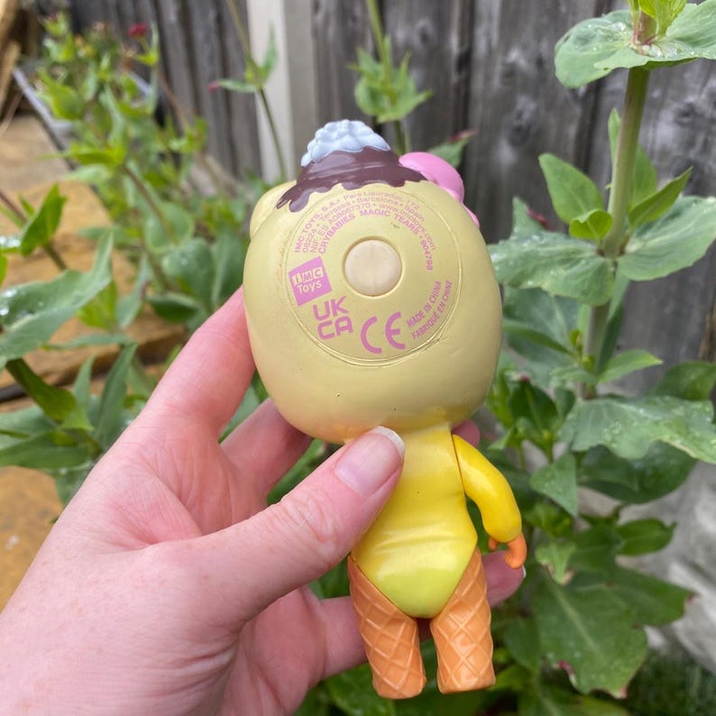 May include: A yellow toy figure with a dessert-themed design. The figure has a light yellow body, orange waffle cone legs, and a head resembling a sundae with brown, pink, and white details. The toy has the text "IMC Toys" and "UK CA CE" printed on the back.