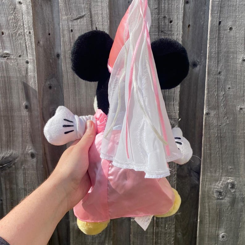 May include: A plush toy featuring a black mouse character wearing a pink satin dress and a matching pink cone-shaped hat with a long, sheer white veil attached. The character has white gloves with black stitched details and yellow feet.