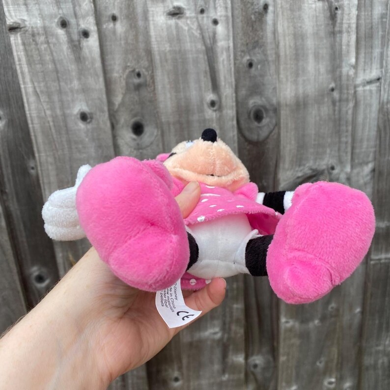 May include: A plush Minnie Mouse doll, featuring a pink dress with white polka dots, large pink feet, and white gloves. The doll has a black nose and ears, and is held against a wooden fence background.